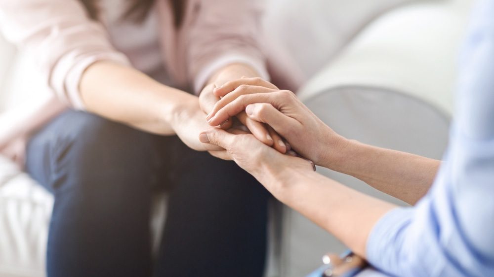 Find Your Inner Strength: Therapy in St. Thomas. One woman offers comfort and reassurance by holding another woman hand during a heartfelt conversation in a cozy living room setting, cropped.